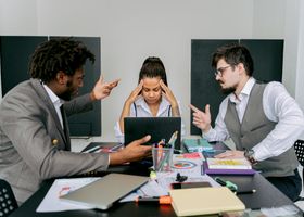 Two men argue while a woman looks frustrated at a laptop in an office environment.