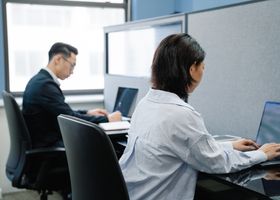 Two professionals working on laptops in a modern office cubicle setup.